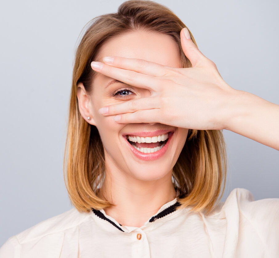 Blond young woman with beaming smile is looking through her hand. She is wearing casual outfit and stands on light grey background